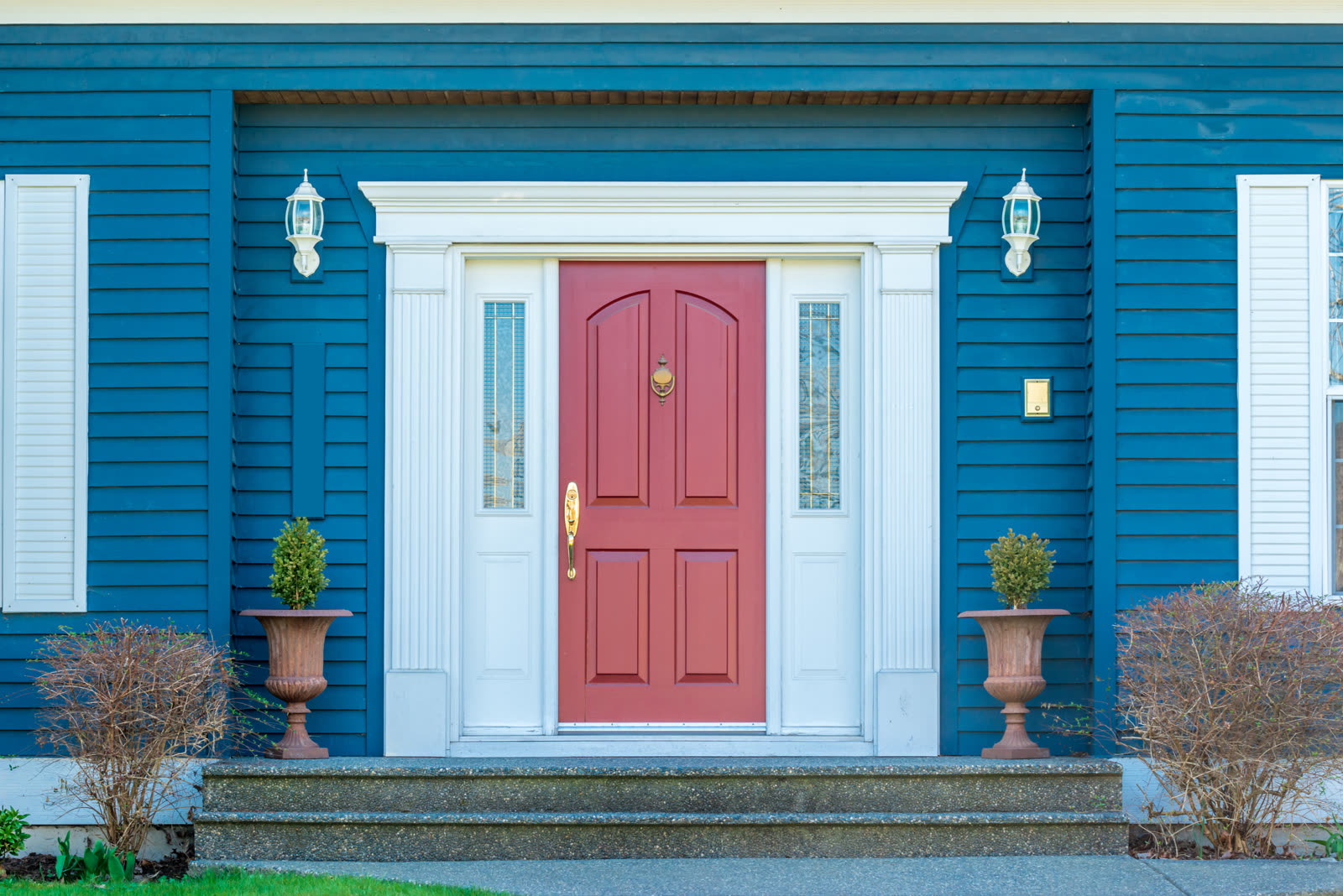 2026-09-are-fiberglass-entry-doors-good-hurricane-zones-sc-2026 A red front door flanked by two narrow glass sidelights and carriage lanterns in the white-trimmed entry of a blue clapboard house with white shutters.